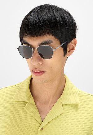 Giovane uomo con capelli neri corti che indossa occhiali da sole ottagonali scuri e una camicia giallo chiaro con trama e colletto, guarda leggermente verso il basso.