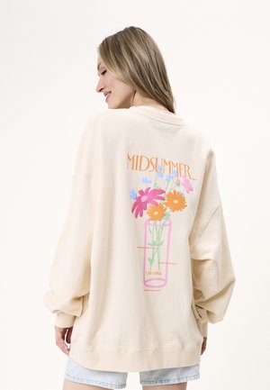Kvinde med langt blond hår har en oversized cremet sweatshirt på med farverigt blomsterprint og teksten "MIDSUMMER" på ryggen.