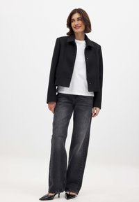 Schwarze cropped Jacke mit einem Kragen und fünf Knöpfen, getragen über einem weißen T-Shirt, kombiniert mit hochgeschnittenen grauen Jeans und schwarzen High Heels.