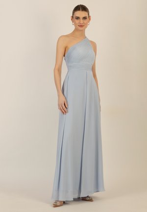 Junge Frau, stehend, trägt ein hellblaues, einseitig schulterfreies Abendkleid mit plissiertem Oberteil, bodenlangem Rock und dekorativen Schuhen.