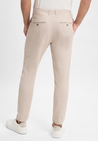 Lichtbeige smart-casual broeken met een slim fit, achterzakken met knopen en een gladde textuur, gecombineerd met lichtgekleurde sneakers.
