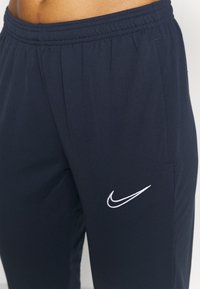 Tmavě modré kalhoty Nike s elastickým pasem, bočními kapsami a bílým vyšívaným logem na levé nohavici. Hladká textury látky.