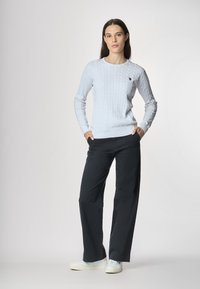 Maglione azzurro chiaro a maglia grossa con maniche lunghe, abbinato a pantaloni neri a gamba larga e sneakers bianche. Il maglione presenta un piccolo emblema.