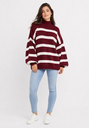 Woman wearing an oversized maroon and white striped sweater, light blue skinny jeans, and white sneakers standing against a plain background.