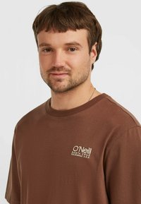 Brun bomulds T-shirt med korte ærmer, rund halsudskæring og et lille hvidt broderet logo på venstre bryst. Glat tekstur.