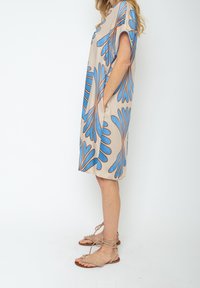 Vestido beige con estampado de hojas azules, mangas cortas y bolsillos laterales. Combinado con sandalias beige con cordones. Tejido suave con un ligero brillo.