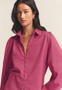 Fitted, deep pink blouse with a collar, decorative button placket, and puffed sleeves made from a lightweight, textured fabric.
