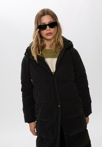 Sort pufferjakke med hætte, lynlåslommer og en tætsiddende design. Laget over en grøn og creme-stribet sweater.