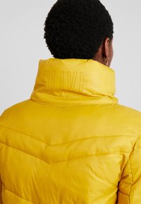 Personne aux cheveux courts et bouclés portant une doudoune jaune vif avec "S. Oliver" gravé sur le col montant, vue de dos.