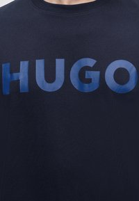 Μπλούζα σε ναυτικό μπλε με στρογγυλή λαιμόκοψη και μεγάλα, λαμπερά μπλε γράμματα που σχηματίζουν τη λέξη "HUGO" στο στήθος.