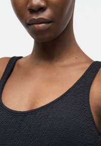 Κοντινό πλάνο ενός ατόμου που φοράει υφασμάτινο μαύρο αμάνικο, δείχνοντας τον λαιμό, τους ώμους και μέρος του προσώπου με ουδέτερη έκφραση.