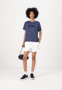 Marineblå bomulds t-shirt med "GANT" print, hvide shorts, sorte sneakers og en lille sort taske. Modellen står foran en hvid baggrund.