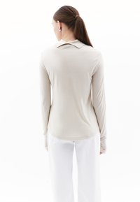 Beige langemou shirt met een kraag, aansluitend ontwerp en zachte textuur, gecombineerd met witte wijde broek, gezien vanaf de achterkant.