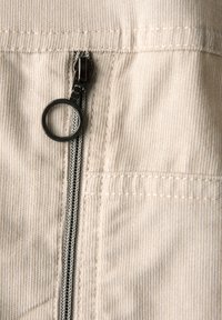 Beige kangas, jossa on hieno nauhatekstuuri, keskisaumassa on suljettu vetoketju mustalla vetotapilla ja hopeisilla hampailla.