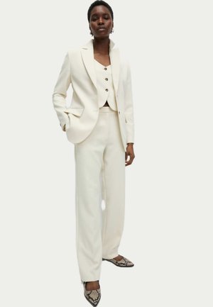 Ivory three-piece suit with a tailored blazer, fitted vest featuring buttons, and wide-leg trousers; accented with snakeskin-patterned heels.