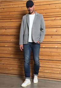 Harmaa bleiseri, jossa on kaksi etutaskua, valkoisen t-paidan päällä, yhdistettynä sinisiin farkkuihin ja valkoisiin lenkkareihin, puupanelin taustalla.