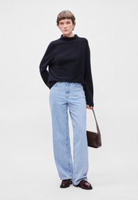 Μαύρος, φαρδύς πουλόβερ με μανίκια, ανοιχτό μπλε, ψηλόμεσα τζιν, καφέ τσάντα; μοντέλο που στέκεται με τα χέρια στις τσέπες, με ίσιες γραμμές και γυαλιστερά παπούτσια.