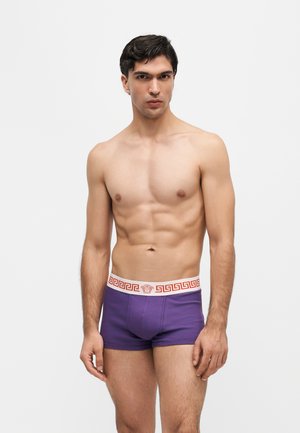 Homme torse nu aux cheveux courts et foncés portant un boxer violet avec une ceinture blanche et rouge à motif sur un fond blanc uni.