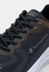 Black athletic shoe with a textured fabric upper, orange accents, and a white sole. Features rounded toe and lacing system for secure fit.