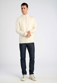 Krämfärgad zip-up sweatshirt med långa ärmar, kombinerad med mörkblå denimjeans och vita sneakers. Enkel design, inga synliga mönster.