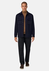 Marineblå knappad jacka med två bröstfickor, bärs över en brun polotröja, i kombination med mörka jeansbyxor och bruna slip-on skor.