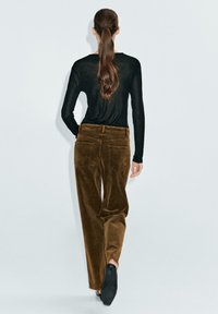 Pantalon en velours côtelé marron avec poches, porté avec un haut noir à manches longues. Les cheveux du modèle sont en queue de cheval basse, montrant la vue arrière.