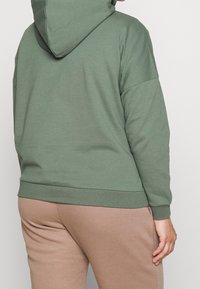 Sudadera verde con capucha hecha de tela suave, que presenta un ajuste relajado y puños acanalados. Usada con pantalones deportivos marrón claro.
