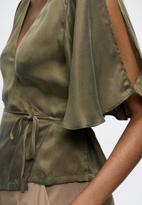 Haut portefeuille en satin vert olive avec manches courtes volantées et découpes latérales, noué à la taille, porté avec un pantalon beige.