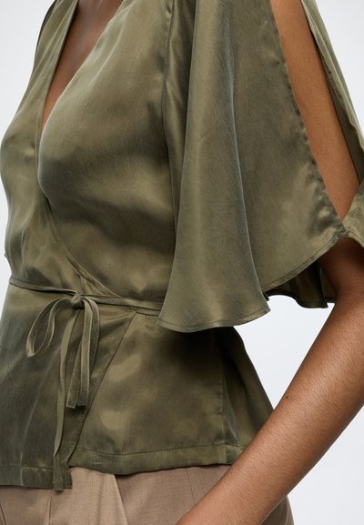 Haut portefeuille en satin vert olive avec manches courtes volantées et découpes latérales, noué à la taille, porté avec un pantalon beige.