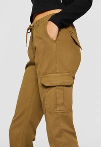 Pantalones cargo de color marrón con cinta elástica en la cintura y cordón. Incluye bolsillos laterales y un bolsillo de solapa en el muslo. Textura de tela suave.