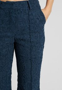 Pantalones azul marino hechos de tela de encaje texturizado, con siluetas ajustadas, bolsillos laterales y una cintura suave con costuras mínimas.