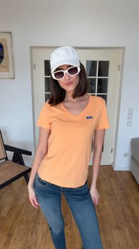 Γυναίκα με σκούρα μαλλιά μέχρι τους ώμους, που φοράει λευκό καπέλο, λευκά γυαλιά ηλίου, πορτοκαλί μπλούζα Hugo και μπλε τζιν σε ένα δωμάτιο με ξύλινο πάτωμα.