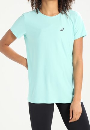 Frau trägt hellblaues kurzärmeliges Sportshirt mit schwarzem Logo auf der Brust, kombiniert mit schwarzen Leggings, steht mit einer Hand auf der Hüfte.
