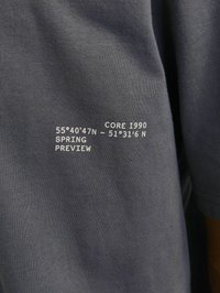 T-shirt en coton gris avec du texte imprimé blanc, incluant des coordonnées et "CORE 1990 SPRING PREVIEW". Texture lisse avec des manches courtes.