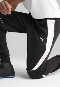 Crne trenirke s bijelim bočnim trakama, s patentnim zatvaračem i Puma logotipom. Materijal izgleda teksturirano s prilagođenim dizajnom.