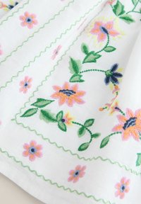 Fehér textile virágmintázat pink, őszibarack és kék hímzéssel, zöld levelekkel és cikk-cakk díszítéssel a széleken.