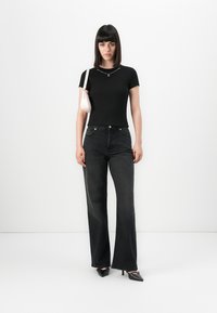 Woman with straight dark hair wearing black fitted t-shirt, high-waisted black wide-leg jeans, black heels, and carrying a small white shoulder bag.