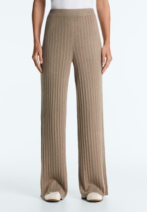 Bézs színű, bőszárú, bordázott kötött nadrág fehér lapos cipővel, deréktól lábfejig egy egyszerű, világos háttér előtt.