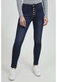 Jeans de tiro alto azul oscuro con un cierre de cinco botones, ajuste slim y un ligero desvanecimiento. Combinados con zapatillas blancas y una camiseta de rayas.