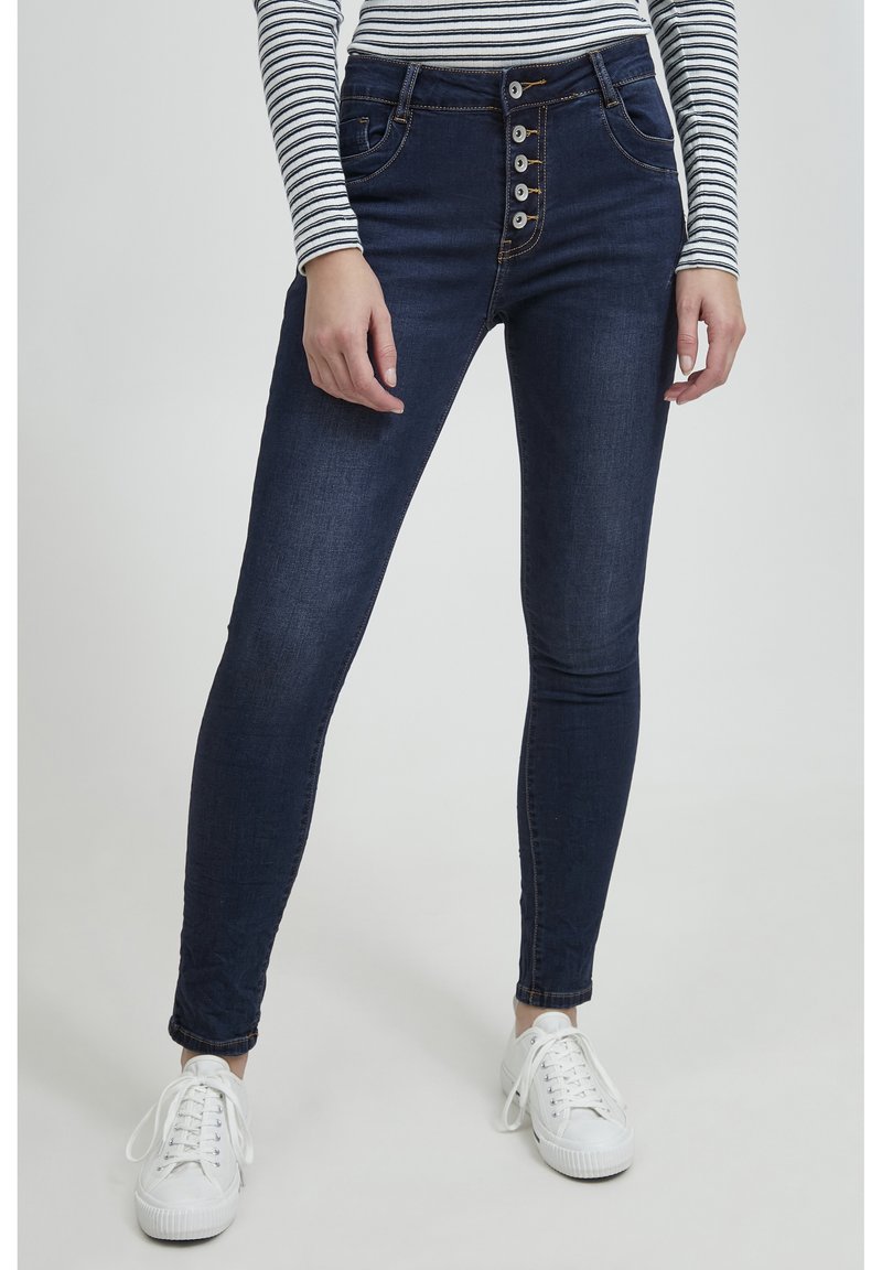 Jeans de tiro alto azul oscuro con un cierre de cinco botones, ajuste slim y un ligero desvanecimiento. Combinados con zapatillas blancas y una camiseta de rayas.