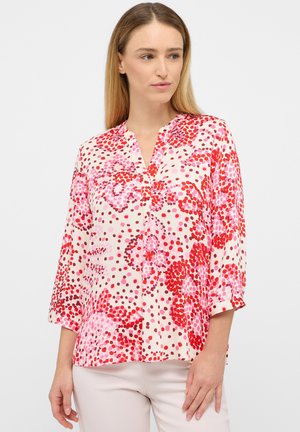 Vrouw die een blouse met lange mouwen draagt met rode en roze abstracte stippenpatronen, gecombineerd met lichtgekleurde broek, staand tegen een effen achtergrond.