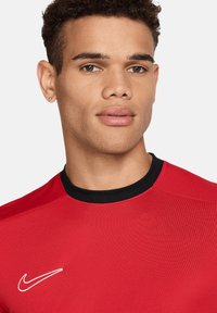 Maglietta rossa Nike con colletto a girocollo nero, caratterizzata da un logo Nike bianco sul lato sinistro. Tessuto liscio con maniche corte.