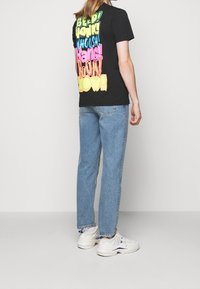 Sort T-shirt med farverig tekst på ryggen i grøn, blå, pink og gul. Parret med lysblå denimjeans og hvide sneakers.