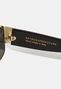Lunettes de soleil avec des montures en plastique noir, des accents dorés, des verres foncés, et un texte imprimé sur la branche indiquant "RETROSUPERFUTURE Fabriqué à la main en Italie."