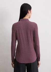 Blouse bordeaux à manches longues avec un motif géométrique blanc, un col et un ourlet arrondi. Tissu à texture lisse. Vue de dos montrée.