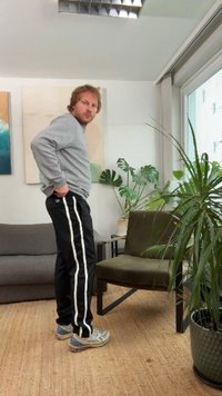 Siva jopa, črne hlače z belimi stranskimi črtami in sivi športni copati. Notranja postavitev z rastlinami in sivim kavčem v ozadju.