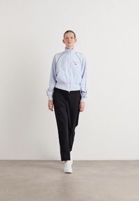 Lichtblauwe ritsjack met zwarte accenten, gecombineerd met zwarte broek en witte sneakers, tegen een neutrale achtergrond.