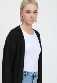 Schwarze Strickjacke mit offenem Vorderteil, die eine leicht strukturierte Oberfläche und subtile metallische Sprenkel aufweist, getragen über einem weißen T-Shirt und einer hellen Jeans.