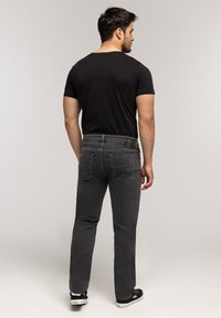 Mężczyzna stojący tyłem, ubrany w czarną koszulkę, ciemnoszare dżinsy i czarne trampki na jednolitym jasnym tle.