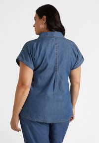 Camisa de denim en lavado medio, con mangas enrolladas, cuello y detalle de pliegue en la parte posterior. Corte recto con dobladillo cosido y costuras laterales.